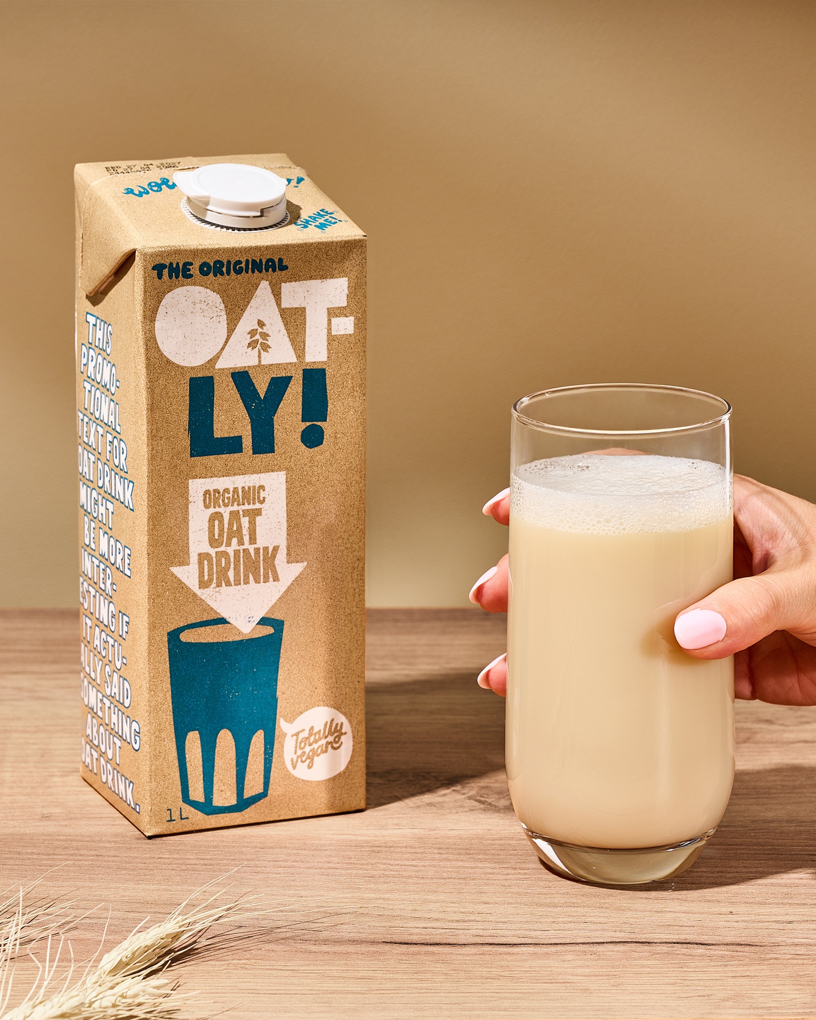 Oatly organic oat drink carton beside a hand holding a glass of oat drink on a warm beige set for Baransu