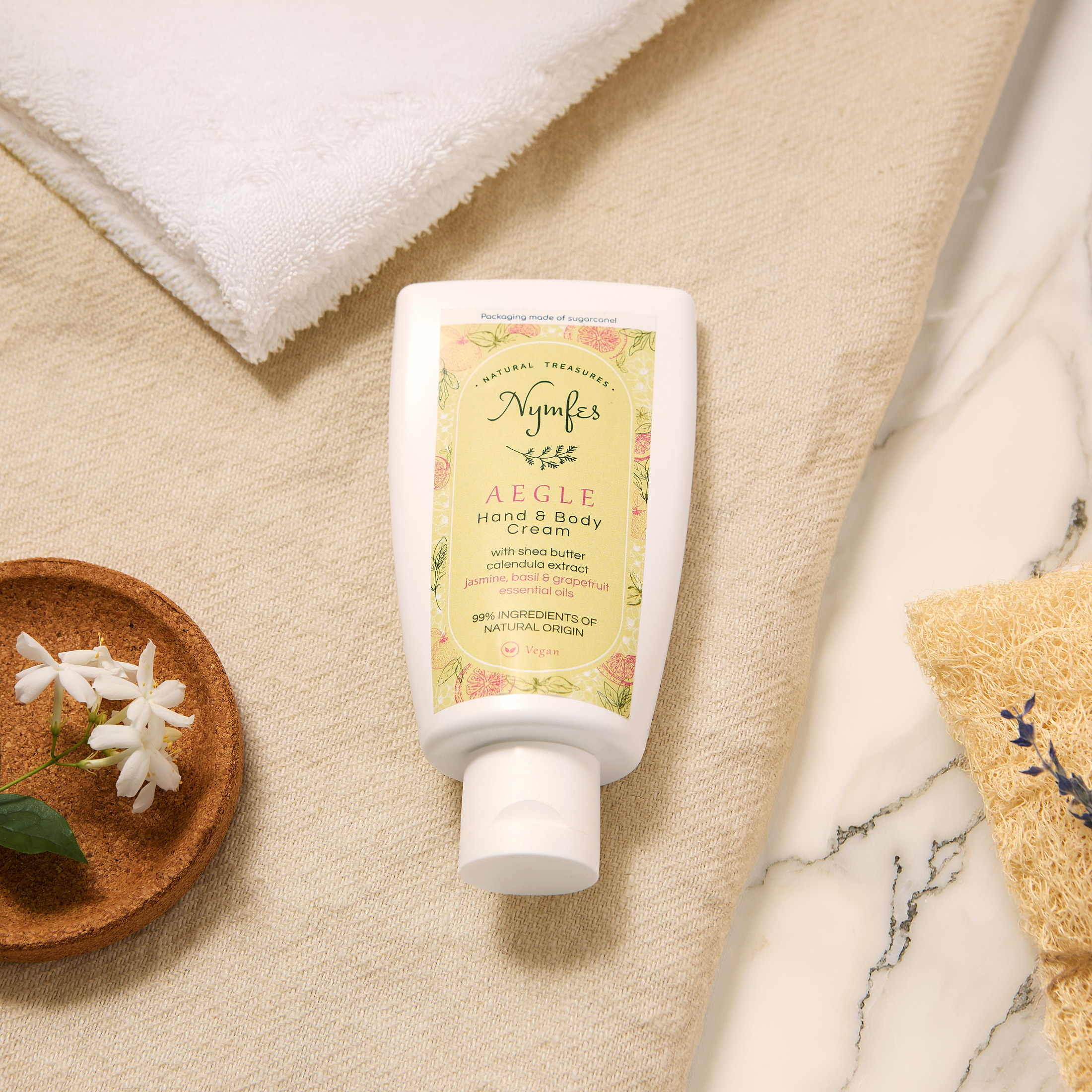 Nymfes Aegle hand and body cream on beige fabric beside jasmine flowers, marble and loofah