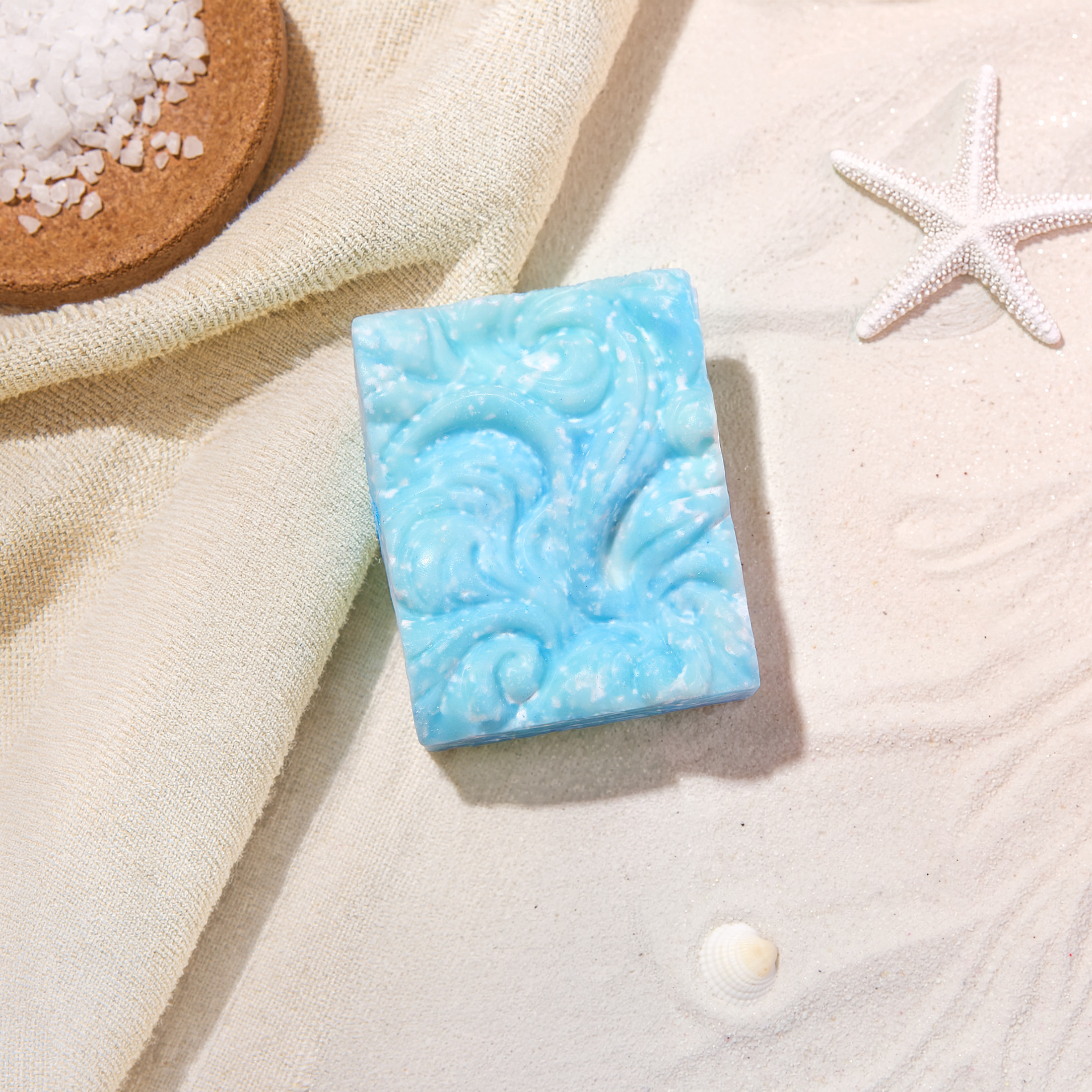 Blue Nymfes soap bar with embossed swirls beside a towel, sea salt and a starfish