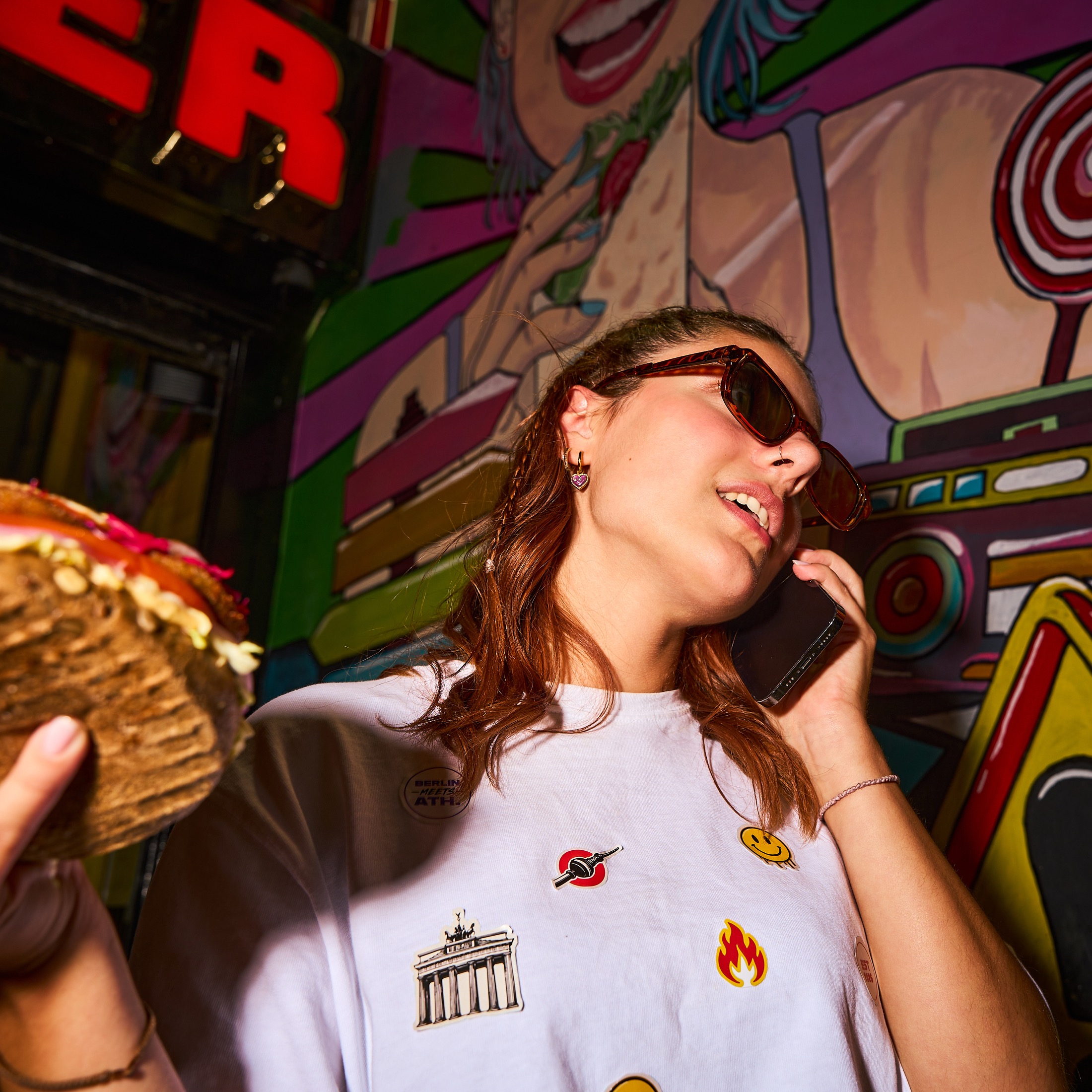 Woman wearing sunglasses, holding a sandwich and talking on a phone outside Super Berlin Doner